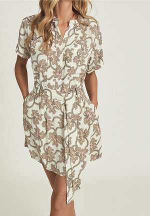 Frau trägt ein kurzärmeliges, tailliertes Kleid mit beigem und braunem Blumen- und Wirbelmuster und steht mit den Händen in den Taschen.