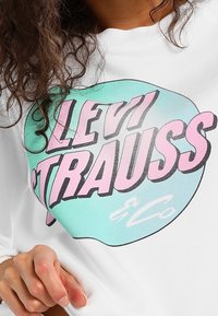 Sudadera blanca con un gran gráfico en texto verde menta y rosa que dice "LEVI STRAUSS & Co." con una forma redondeada. Textura de tela suave.