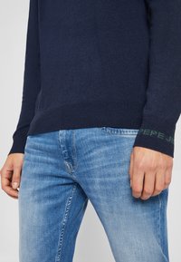 Homme portant un pull en maille bleu foncé avec "PEPE JEANS" sur le poignet et un jean bleu clair délavé, debout avec les mains détendues le long du corps.