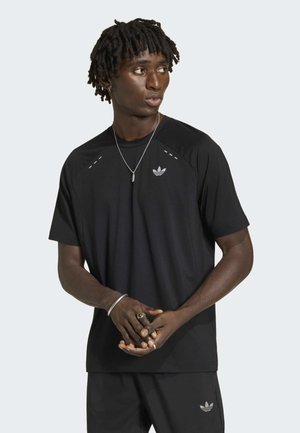 Mężczyzna z krótkimi dredami w czarnej koszulce i szortach Adidas, patrzący na bok, ze złączonymi rękami przed sobą.