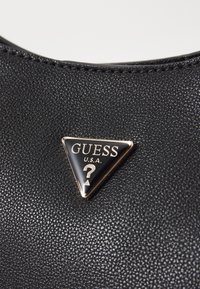 Sort tekstureret materiale med et trekantet metalemblem, der har guldtrim og mærkenavnet "Guess U.S.A." over et spørgsmålstegn.