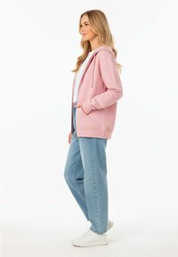 Felpa rosa chiaro con tasche a marsupio e cordino, abbinata a jeans larghi azzurri e sneakers bianche, mostrata di profilo.