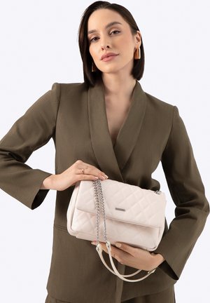 Žena v olivovém obleku držící béžovou prošívanou kabelku se stříbrnými řetízkovými uchy na bílém pozadí.