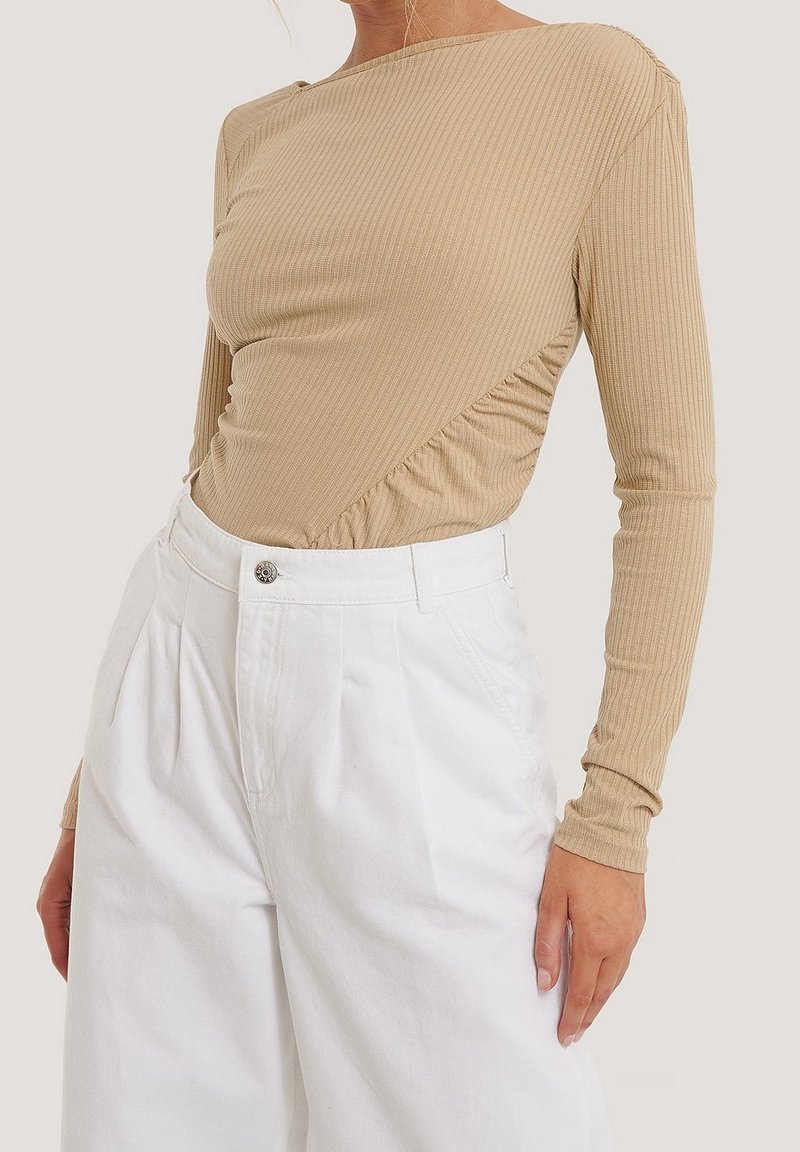 Haut à manches longues côtelé beige clair avec un design asymétrique et des détails froncés sur le côté, associé à un pantalon blanc taille haute.
