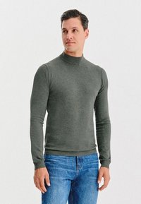 Ciemnozielony sweter z golfem wykonany z miękkiego materiału, z długimi rękawami i prążkowanym wykończeniem u kołnierza i na dole, w zestawie z niebieskimi dżinsami.