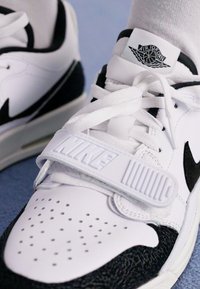 Fehér és fekete kosárlabda tornacipők perforált orr résszel, fűzőkkel és egy pánttal, amelyen a kiemelkedő "NIKE" logó látható az oldalon.