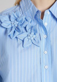 Camisa a rayas de color azul claro con cuello y dos grandes flores de tela en el hombro izquierdo. Presenta botones blancos en el frente.