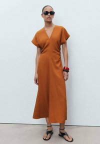 Rust-colored midi dress with a V-neck, short sleeves, and a flowing silhouette. Accessories include a red bangle and black sandals.