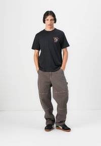 Schwarzes Baumwoll-T-Shirt mit rotem Grafikdruck, kombiniert mit locker sitzenden, braunen, schmal zulaufenden Hosen mit Nähdetails und schwarzen Sneakers.