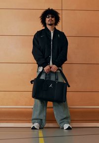 Sort Jordan skuldertaske med dobbelt håndtag, glat tekstur og logo, kombineret med løstsiddende blå jeans og en sort jakke mod en træbaggrund.