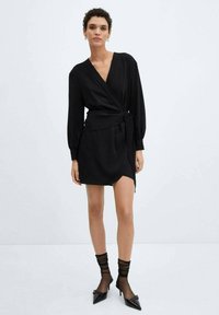 Black wrap dress with long sleeves, featuring a side tie and a short hem. Worn with sheer socks and pointed-toe heels. Smooth texture.