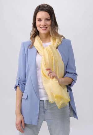 Blazer azzurro chiaro con maniche arrotolate, t-shirt bianca e sciarpa gialla con finitura testurizzata, caratterizzata da orli frangiati. Outfit casual.