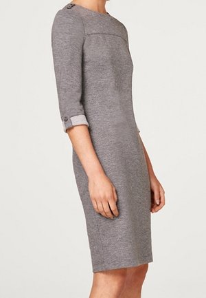 Kobieta w szarej sukience za kolano, dopasowanej, z rękawami do połowy przedramienia i ozdobnymi guzikami na rękawach oraz ramieniu.