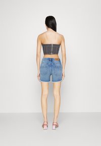 Žena s dlouhými tmavými vlasy stojí zády, má na sobě šedý zipový top bez ramínek, modré džínové šortky a sandály na blokovém podpatku s iridescentním efektem.