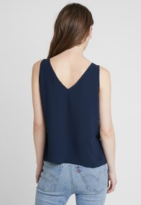 Marinblå ärmlös topp med V-ringning och lös passform, tillverkad i lättviktsmaterial, med rak nederkant och sidoklykor.