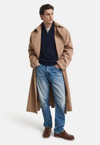 Beige Trenchcoat, marineblauer Pullover, hellblaue Jeans und braune Bootsschuhe. Verfügt über einen Reißverschlusskragen, Taschen und eine entspannte Passform. Einfaches Design.
