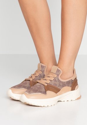 Pieds portant des baskets beige et marron à motifs avec des semelles blanches épaisses sur un sol gris clair.