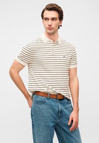 Μπεζ ριγέ πόλο μπλουζάκι με κοντά μανίκια, γιακά και μικρό λογότυπο στο στήθος, σε συνδυασμό με ανοιχτό μπλε τζιν και καφέ ζώνη.