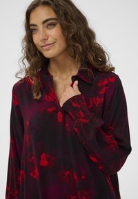 Koszula z długim rękawem z czarnego materiału z czerwonymi abstrakcyjnymi wzorami, z kołnierzykiem, zapinana na guziki z marszczonymi mankietami.