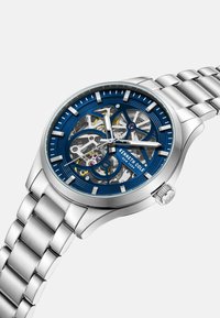 Zegarek skeleton z niebieską tarczą, stalowosrebrną kopertą i bransoletą, złożony z misternych wewnętrznych mechanizmów i świecących znaczników godzin.