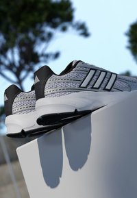 Sivé a čierne atletické topánky s textúrovaným sieťovinovým zvrškom, tromi čiernymi pruhmi a Cloudfoam odpružením na bielom podstavci.