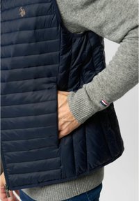 Gilet imbottito blu navy con design a righe verticali, tasca con zip sul lato e un maglione grigio visibile sotto con polsini a coste.