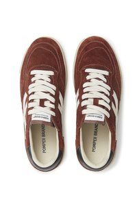 Zapatillas de ante marrón con cordones y rayas blancas, que presentan una punta redondeada y un acento negro en el talón. Detalles de superficie texturizada visibles.