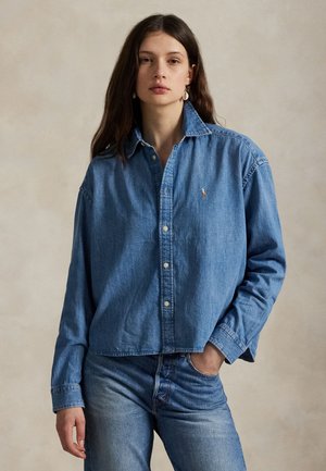 Giovane donna con lunghi capelli scuri indossa una camicia di jeans blu con bottoni e jeans, in piedi davanti a uno sfondo neutro con una mano in tasca.