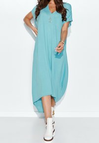 En kortärmad V-ringad klänning i akvamarinblått tyg, med lös passform, asymmetrisk nederkant och slät textur, kombinerad med vita ankelskor.