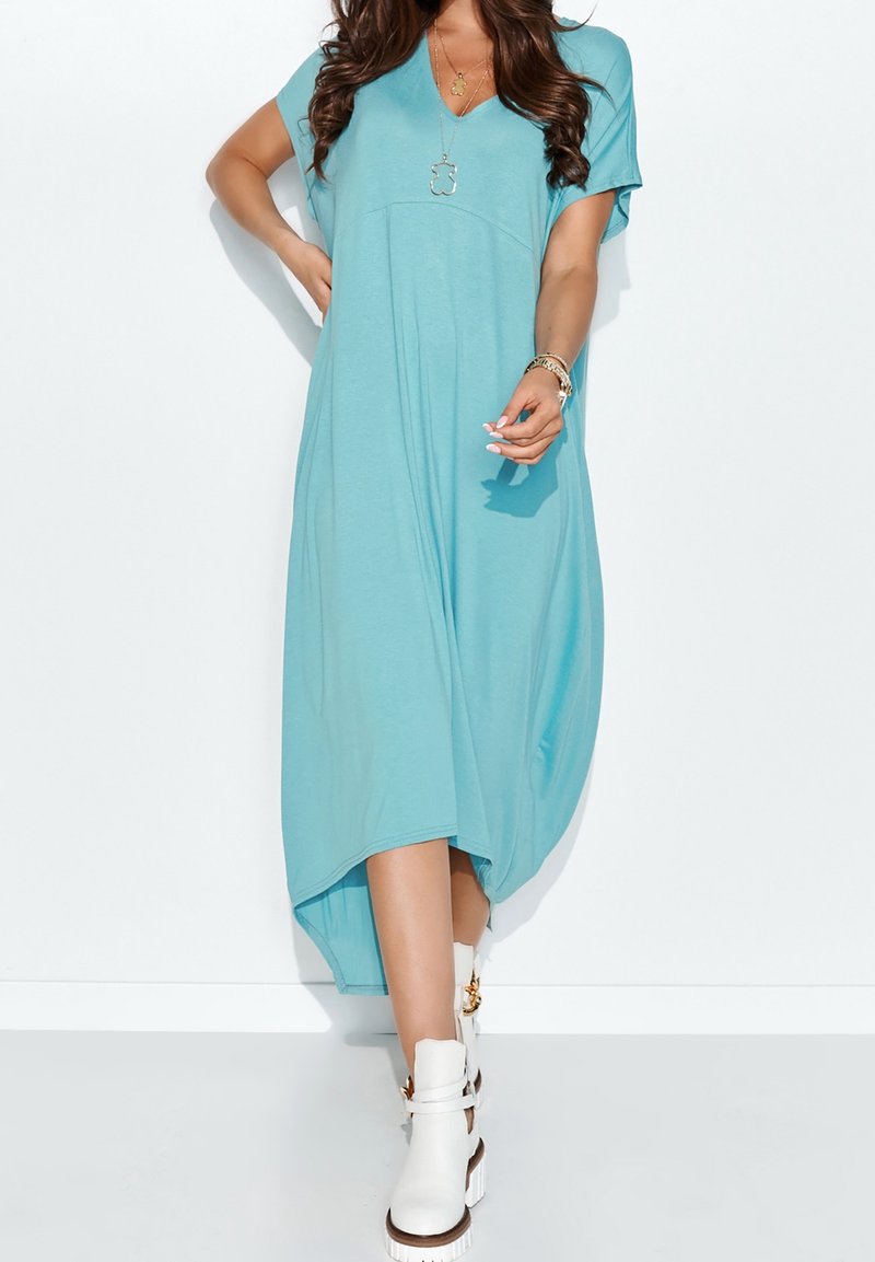 En kortärmad V-ringad klänning i akvamarinblått tyg, med lös passform, asymmetrisk nederkant och slät textur, kombinerad med vita ankelskor.