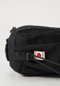 Zaino in tessuto nero con forma arrotondata, chiusura a zip, spallacci regolabili e un distintivo logo rosso e bianco ben visibile.