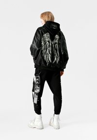 Chaqueta de cuero negro con bordado de alas plateadas, combinada con pantalones negros con estampados gráficos y botas de combate blancas.