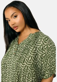 Grön mönstrad blus med en texturerad lövdesign, med en krage och tre knappar. Korta ärmar och en avslappnad passform.