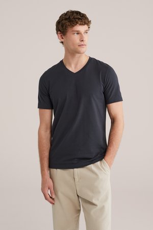 Mladý muž v tmavosivom tričku s V-výstrihom a béžových nohaviciach, stojaci s jednou rukou v vrecku na jednoliatom pozadí.