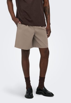 Homme portant un short beige, un t-shirt marron foncé, des chaussettes marron assorties et des mocassins en cuir noir, debout avec une main dans la poche.