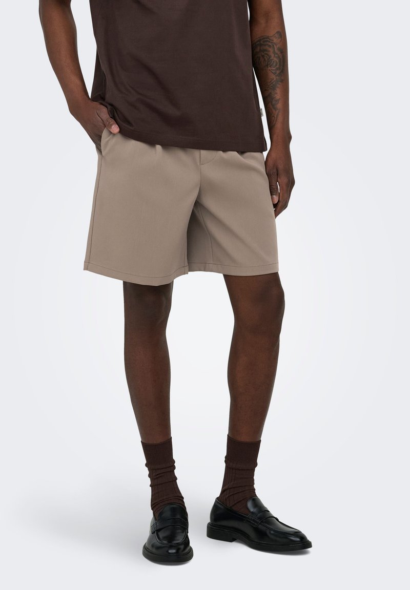 Homme portant un short beige, un t-shirt marron foncé, des chaussettes marron assorties et des mocassins en cuir noir, debout avec une main dans la poche.