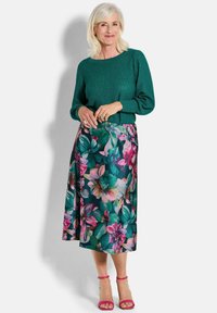 Grüner Strickpullover mit strukturiertem Finish, kombiniert mit einem fließenden, floral-midi Rock, der große rosa und türkisfarbene Blüten zeigt. Rosa hohe Sandalen.