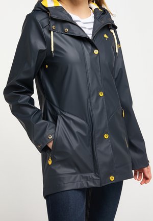 Femme portant une veste de pluie bleu foncé avec des boutons jaunes, des accents jaunes, une capuche et des poches, associée à un jean bleu foncé et une chemise rayée.