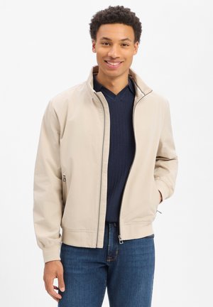Junger Mann lächelt, trägt eine beige Jacke mit Reißverschluss über einem marineblauen Pullover und blauen Jeans, steht vor einem schlichten weißen Hintergrund.