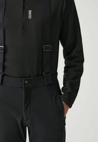 Uomo che indossa pantaloni da sci neri Colmar con bretelle regolabili e una maglietta nera a maniche lunghe, con una mano nella tasca con zip.