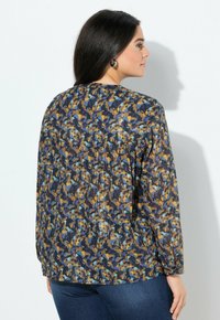 Frau mit langen dunklen Haaren, die eine langärmlige Bluse in Blau, Senfgelb und Petrolmuster und dunkelblaue Jeans trägt, von hinten gezeigt.