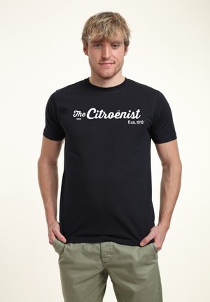 Sort bomulds t-shirt med "The Citroënist" logo i hvid skrift, kortærmet og med rund hals. Båret med lysegrønne bukser.