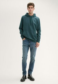 Felpa verde con cappuccio e cordino, tasca a marsupio, polsini a costine; abbinata a jeans blu e stivali neri alla caviglia su uno sfondo neutro.