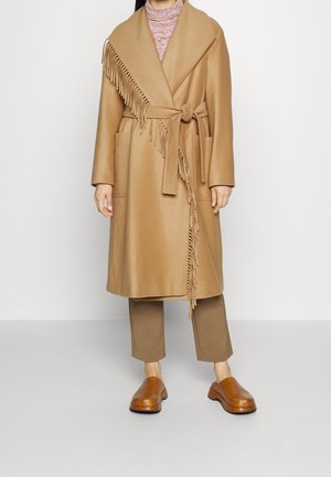 Mujer con abrigo camel con detalle de flecos sobre pantalones marrones y zapatos marrones sin cordones, de pie frente a un fondo neutro.