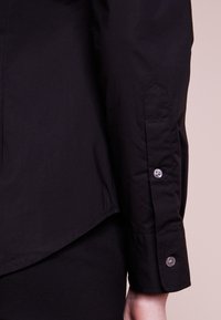 Svart långärmad skjortärm med två knappar, bärs på armen mot en ljusrosa bakgrund.