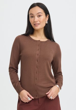 Vrouw met lang donker haar die een bruin gebreid vest met knopen en bijpassende broek draagt, geaccentueerd met zilveren oorbellen, ringen en armband.