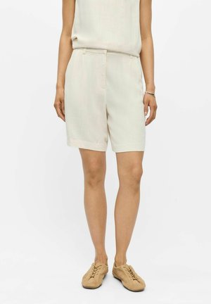 Femme portant un short beige taille haute sur mesure, un top sans manches assorti, des chaussures marron clair à lacets et un bracelet argenté au poignet droit.