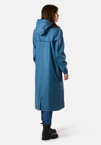 Niebieski wodoodporny długi płaszcz z kapturem, z rozcięciem z tyłu, elastycznymi mankietami i gładką, błyszczącą teksturą. Noszony na dżinsy i botki.