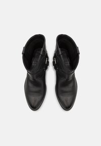 O pereche de cizme negre din piele, cu gleznă, având vârfuri ascutite și curele decorative cu catarame argintii, văzute dintr-o parte de sus pe un fundal alb.