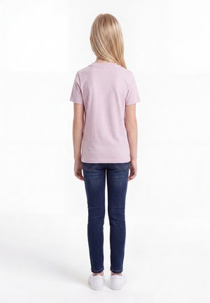 Barn med langt blond hår iført en lyserød kortærmet skjorte, mørkeblå jeans og hvide sneakers, stående med ryggen til på en hvid baggrund.
